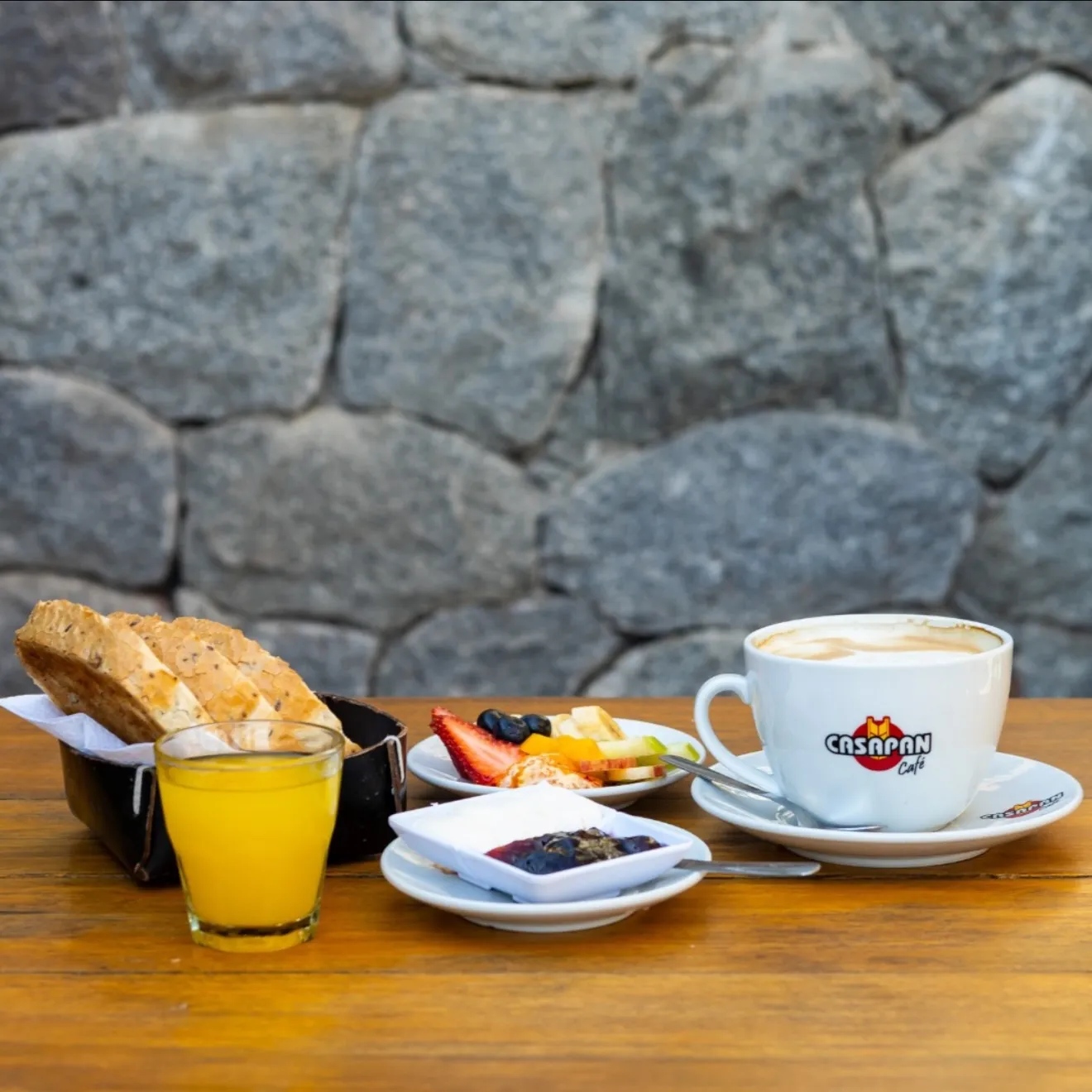Desayuno completo de Casa Pan: tostadas, jugo de naranja, fruta, mermeladas y cappuccino sobre mesa de madera con pared de piedra.