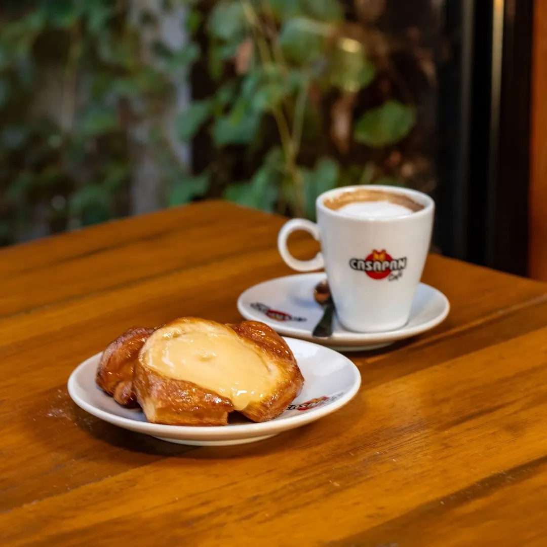 Factura recién horneada rellena con dulce de leche junto a un cappuccino sobre mesa de madera.